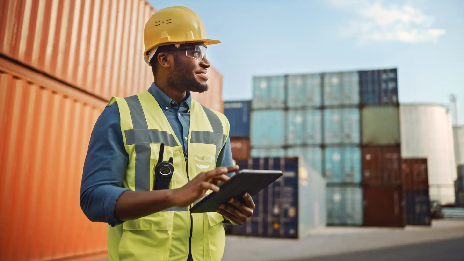Homem trabalhador com capacete de segurança e colete refletivo usa tablet em ambiente de porto de containers, simbolizando gestão de logística e transporte.