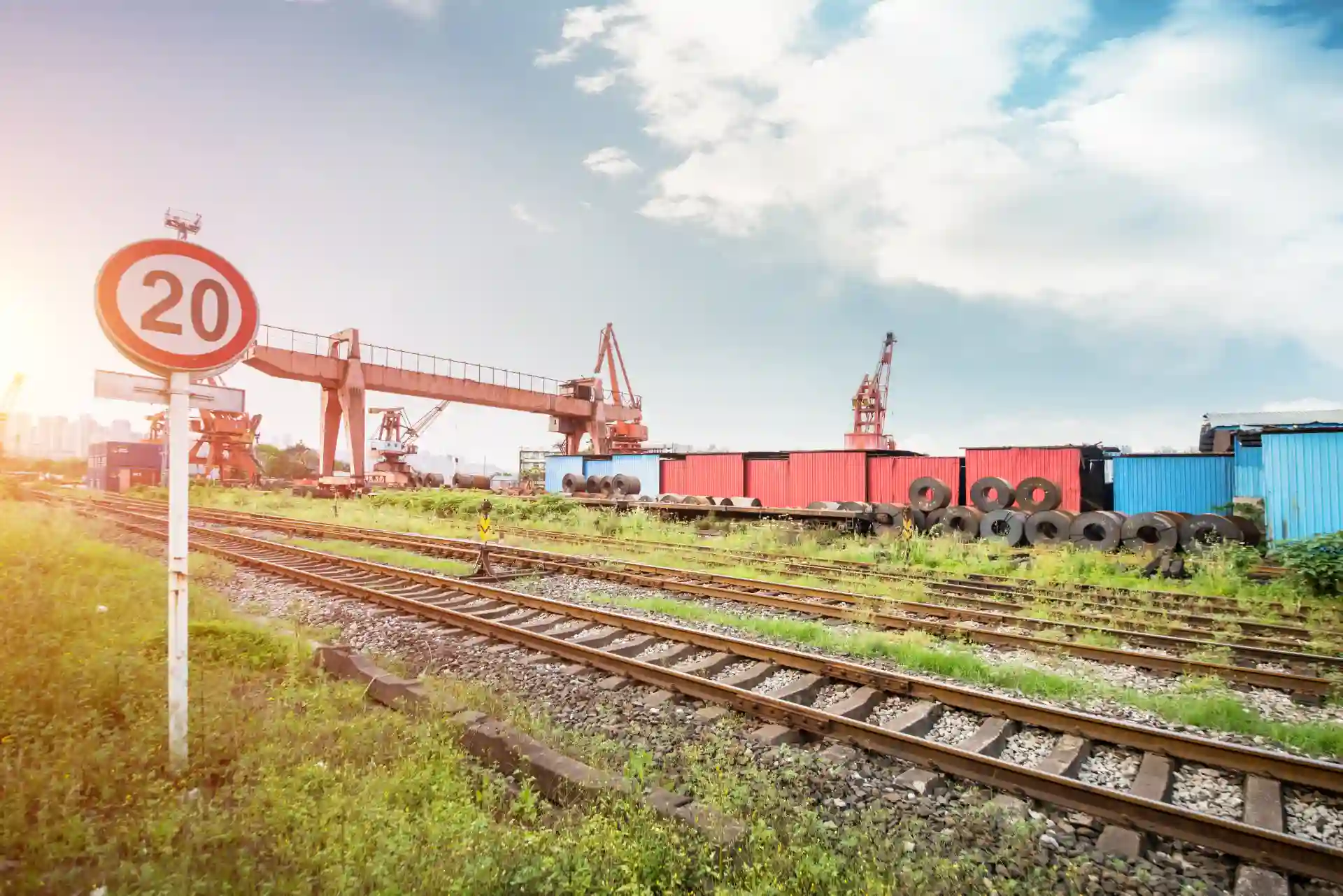 Imagem de uma ferrovia industrial com trilhos, carga, torres de transporte e céu nublado ao fundo, capturada ao entardecer.