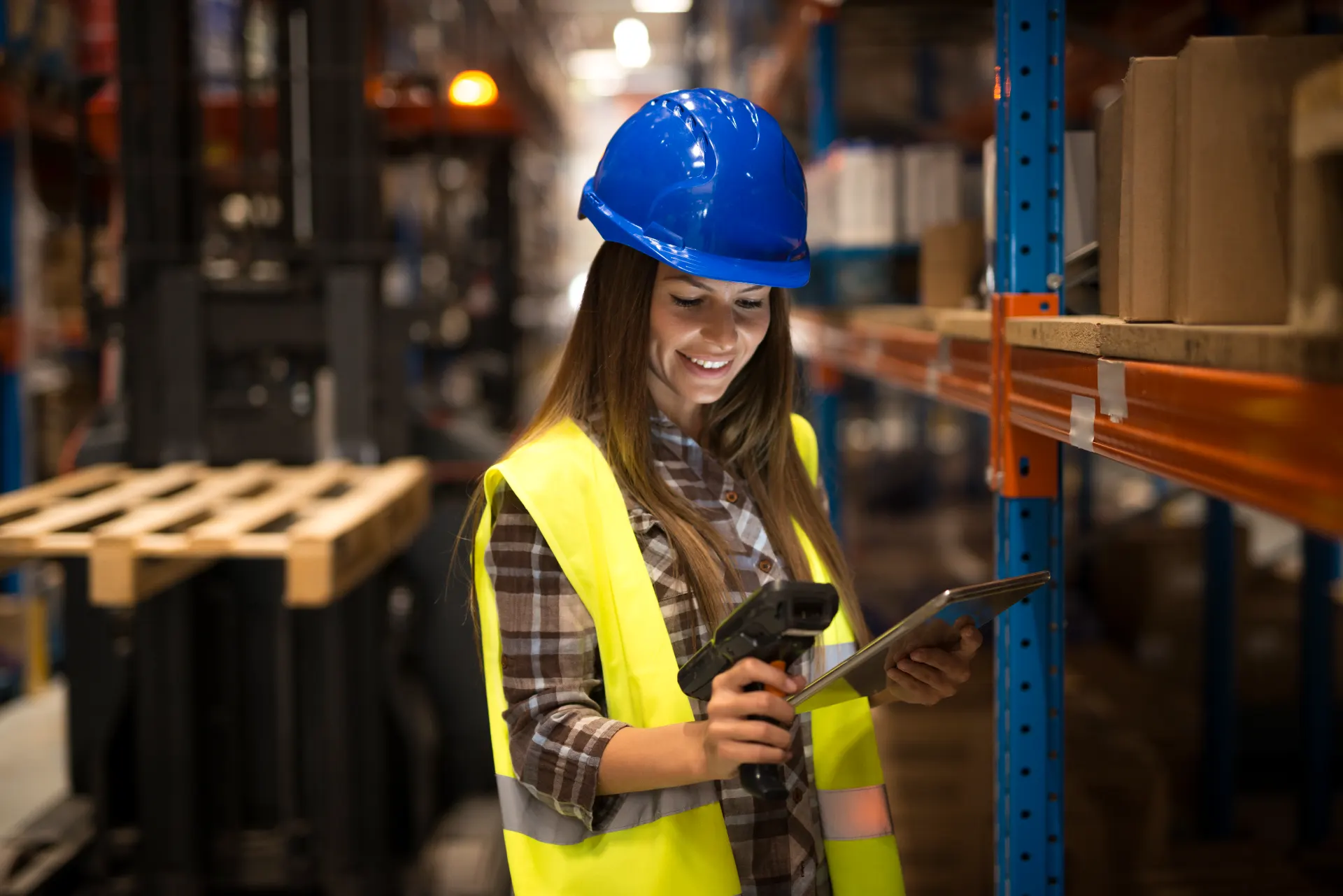 Trabalhadora em armazém logístico usando capacete e colete de segurança, conferindo informações em tablet ao lado de prateleiras com caixas e pallets, relacionada à classificação fiscal na importação.