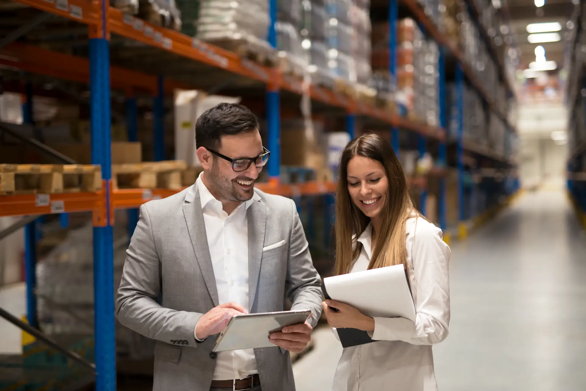 Equipe em um armazém logístico, com caixas e estrutura de prateleiras ao fundo, enquanto um consultor e uma profissional analisam documentos em tablets e pastas. Serviço de assessoria aduaneira e acompanhamento de cargas.