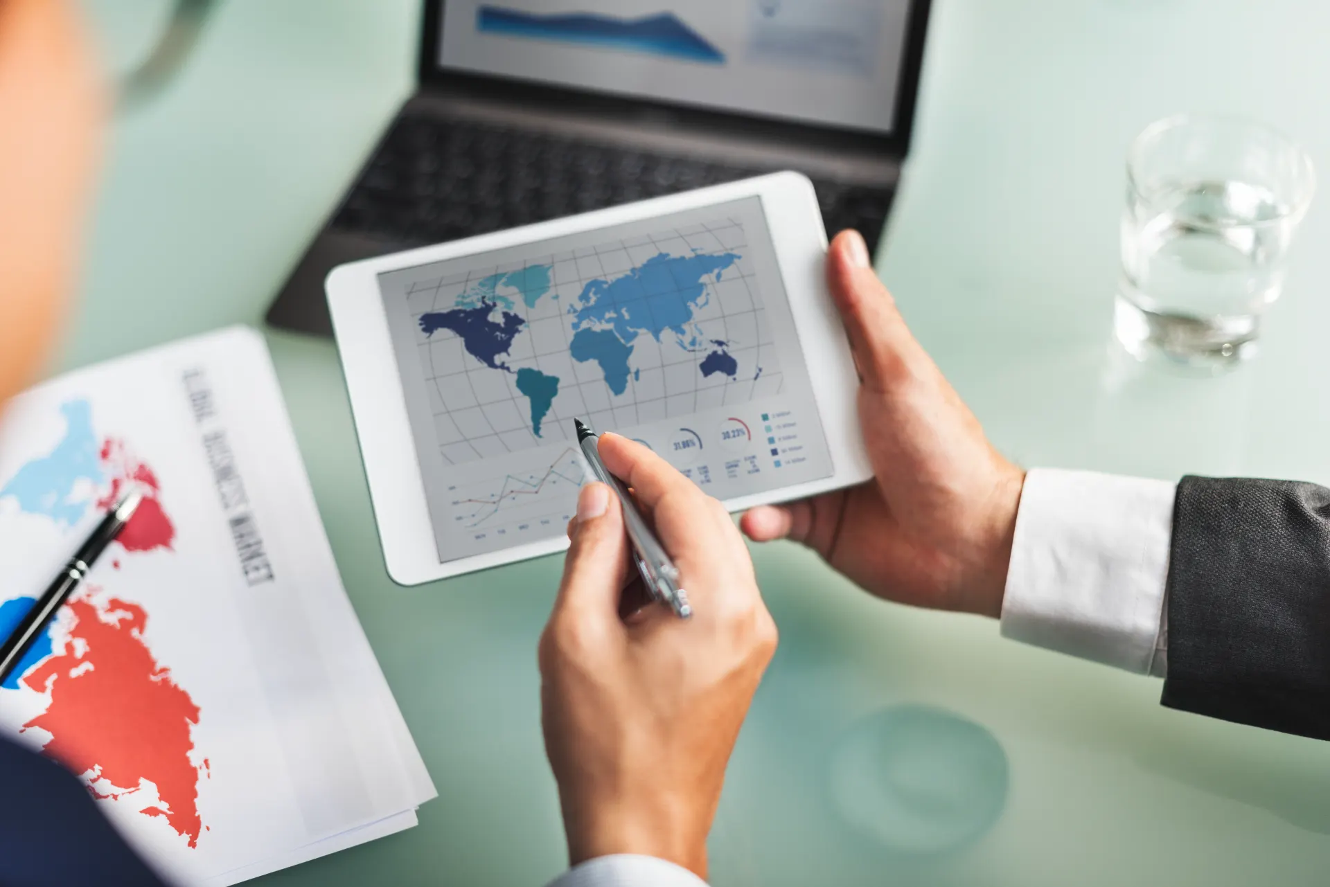 Pessoa analisando mapa-múndi e gráficos em tablet durante reunião de negócios, com documentos e pesquisa para planejar importar do mercosul